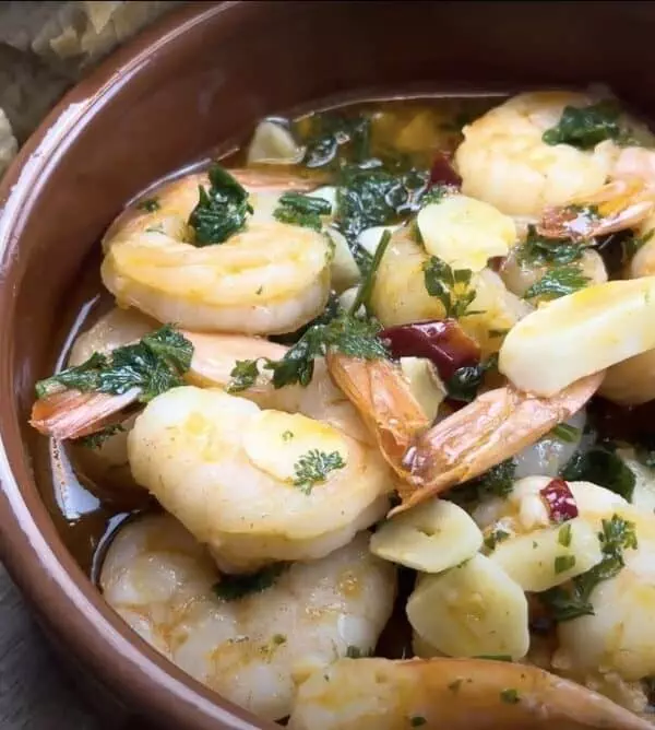 Garlic shrimp tapas in a brown dish with fresh herbs, served alongside crusty bread slices on a wooden table.